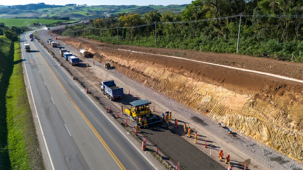 Perspectiva aérea de uma rodovia, com obras na marginal e maquinários trabalhando.