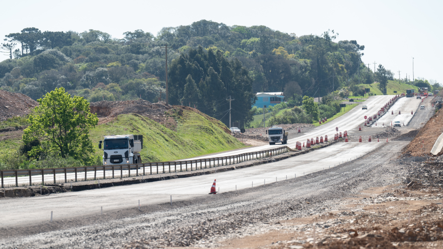 Rodovia em construção, com cones laranjas nas margens da estrada e caminhões trafegando.