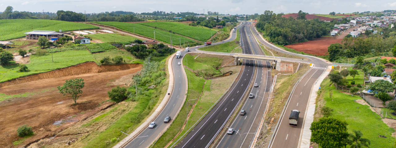 Vista aérea de uma rodovia com múltiplas faixas de tráfego próximas a edifícios, e plantações.