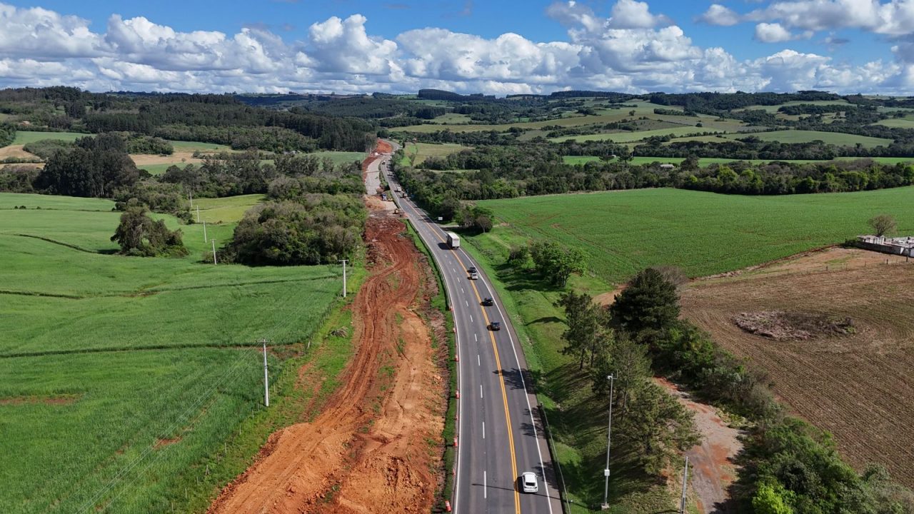 Perspectiva aérea de uma rodovia, com obras na marginal e maquinários trabalhando.