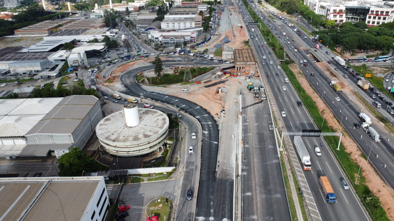 Vista aérea de uma rodovia com múltiplas faixas de tráfego próximas a edifícios industriais e armazéns, com uma paisagem urbana e colinas verdes ao fundo sob um céu parcialmente nublado.