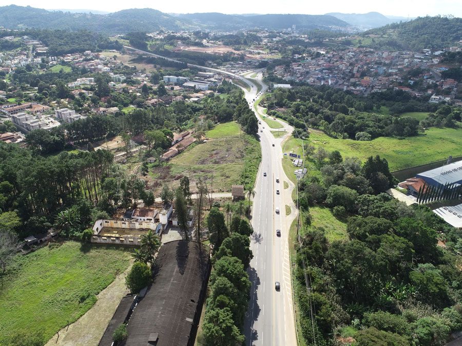 Perspectiva aérea de uma rodovivia em meio à floresta, com obras e a infraestrutura em desenvolvimento as margens da rodovia e residências ao fundo.