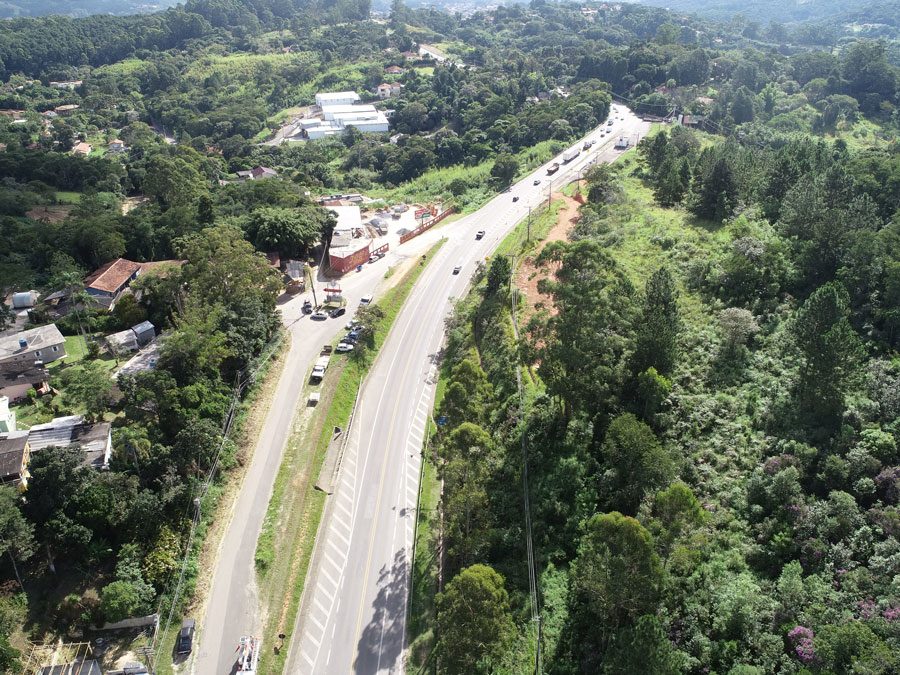 Perspectiva aérea de uma rodovivia em meio à floresta, com obras e a infraestrutura em desenvolvimento as margens da rodovia e residências ao fundo.